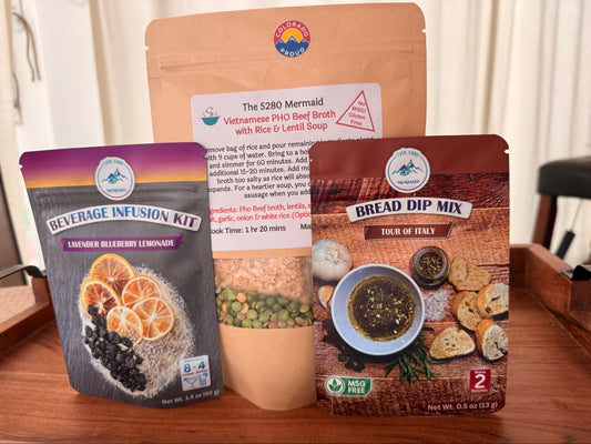 Family Flavor Bundle: Lavender Blueberry Lemonade Beverage Infusion Kit pouch, Vietnamese Pho Beef Broth with Rice and Lentil Soup pouch, and Tour of Italy Bread Dip Mix pouch on a wooden tray